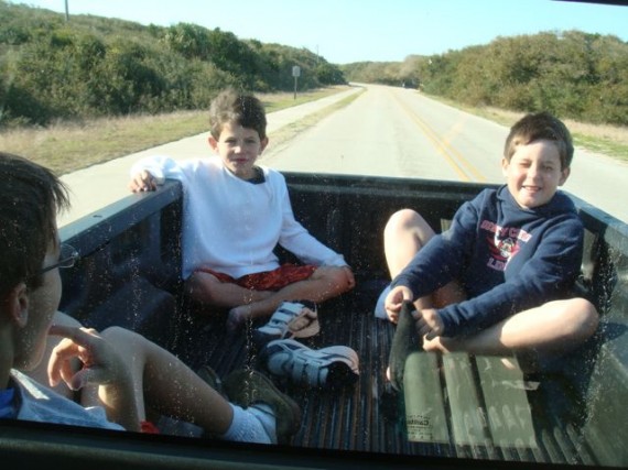 Three young boys in the back of a pickup truck driving down a country road